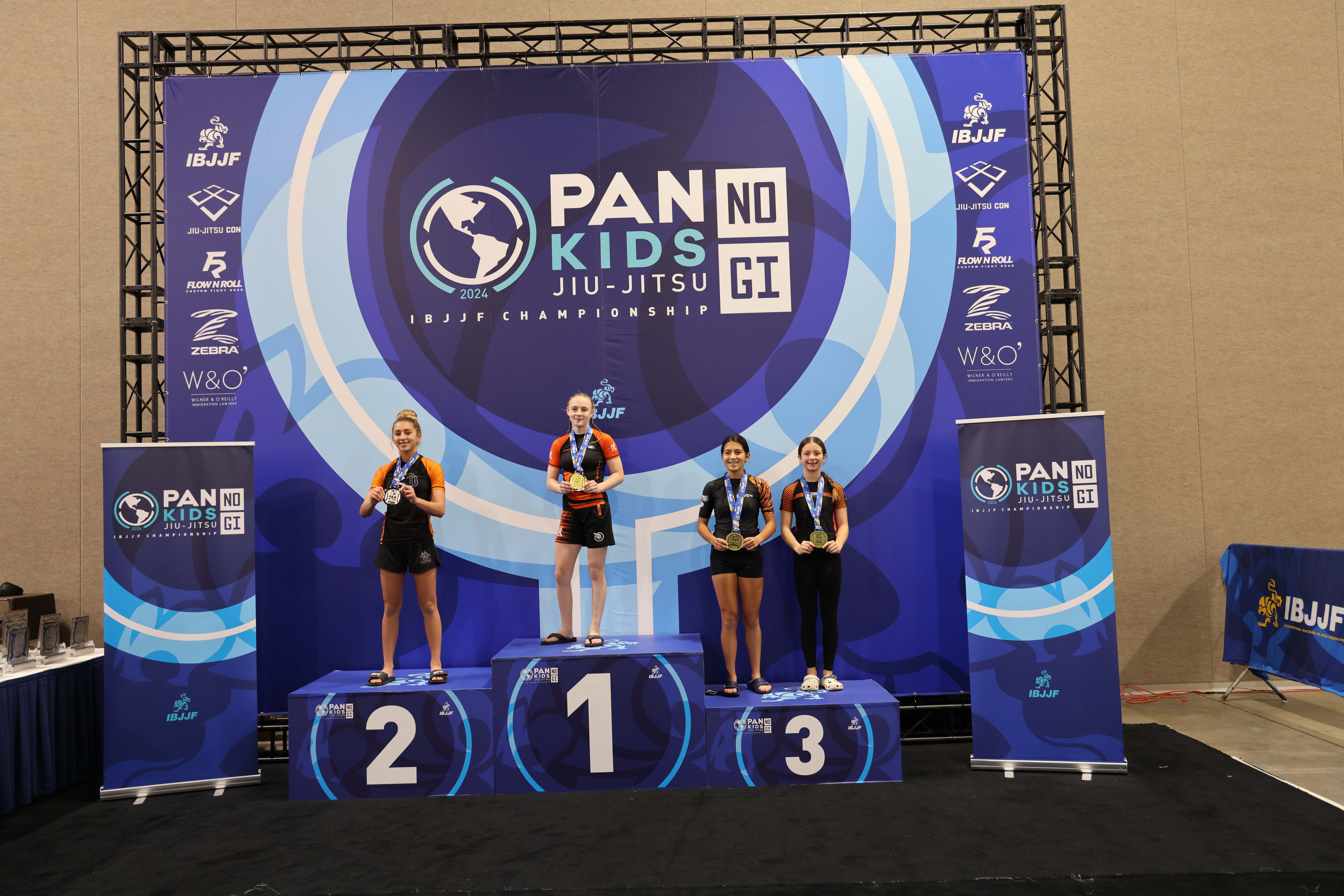 Φωτογραφία βάθρου από το Pan Kids IBJJF No-Gi Championship 2024. Το ψηλότερο σκαλί του βάθρου καταλαμβάνεται από την πρωταθλήτρια της κατηγορίας. Η σήμανση της διοργάνωσης είναι ορατή στο φόντο: PAN KIDS NO-GI IBJJF CHAMPIONSHIP.