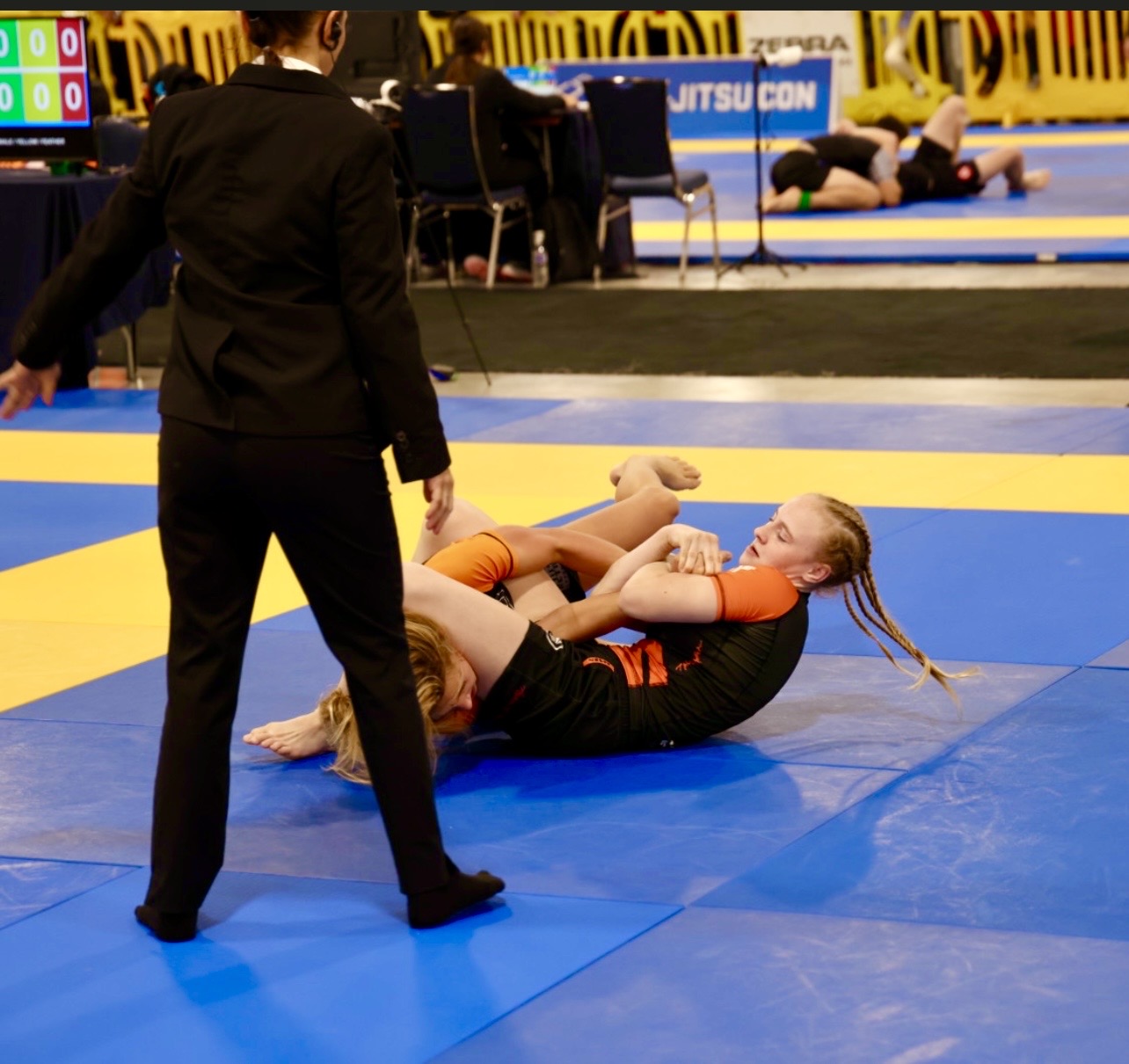 Αγωνιστική εικόνα από το ταμπλό του IBJJF Pan Kids No-Gi Championship 2024. Το υποκείμενο εμφανίζεται σε θέση ελέγχου από πάνω. Αγωνιστικό περιβάλλον υπό την αιγίδα της IBJJF, με το τραπέζι βαθμολόγησης και τα διπλανά στρώματα ορατά στο φόντο.